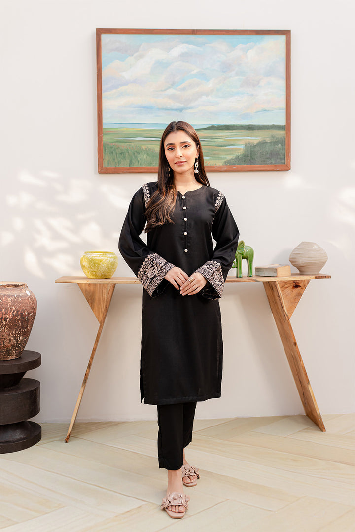 Woman in black embroidered tunic and black pants, with floral pink sandals, in a modern interior.