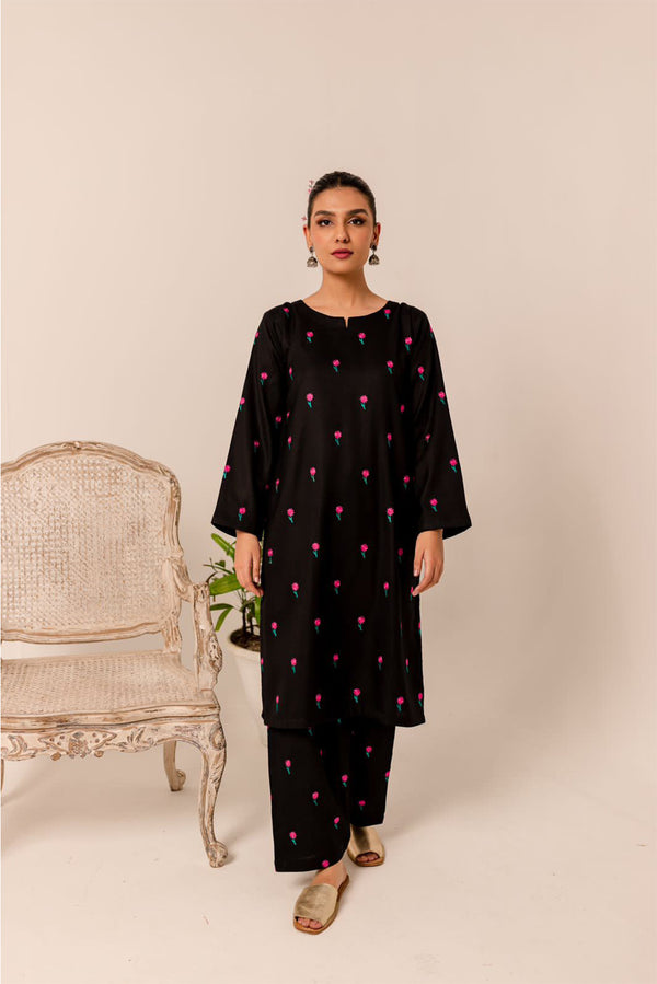 A woman in a black embroidered tunic and matching pants, standing beside a vintage chair and a plant.