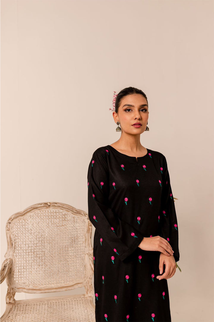 A woman in a black floral-embroidered dress standing beside a woven chair.