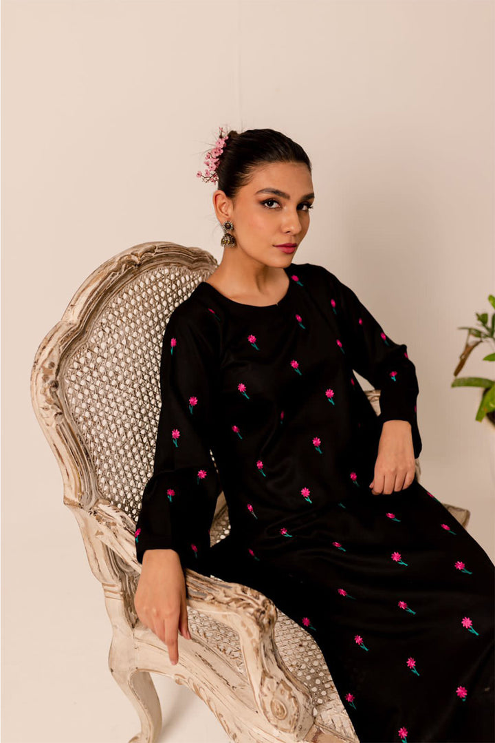 A woman in a black embroidered dress with pink flowers, seated on a vintage white chair.