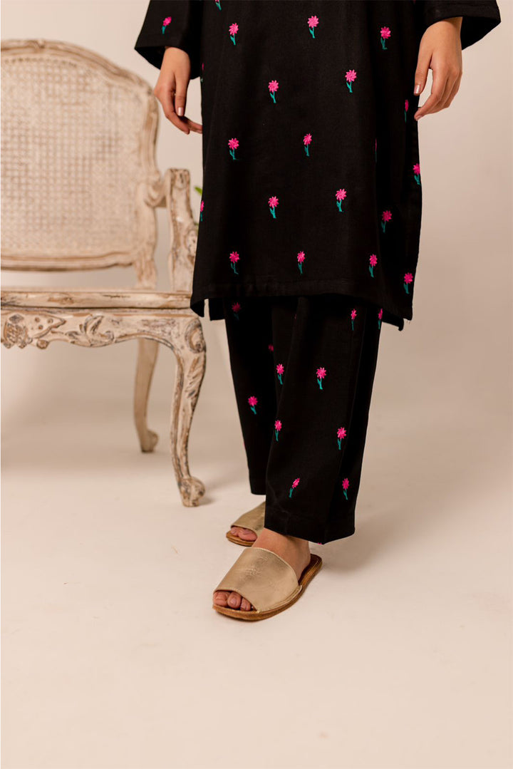 Woman in black embroidered two-piece cotton slub outfit with floral patterns, standing beside a decorative chair.