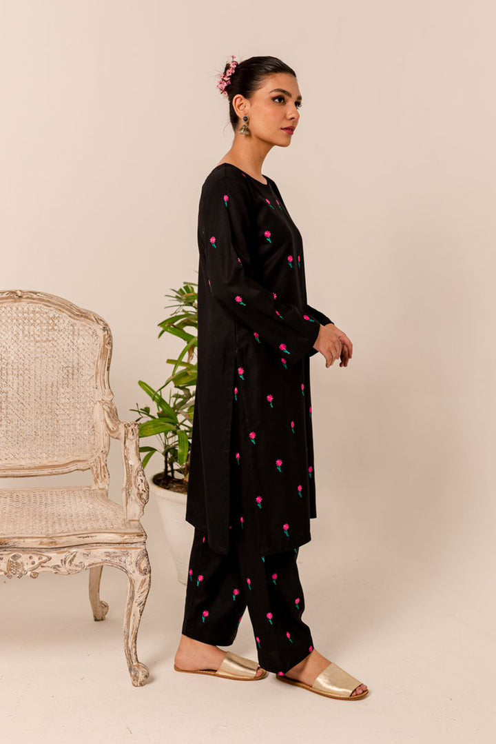 Model in black two-piece outfit with pink floral embroidery, paired with gold sandals, standing next to a wicker chair.